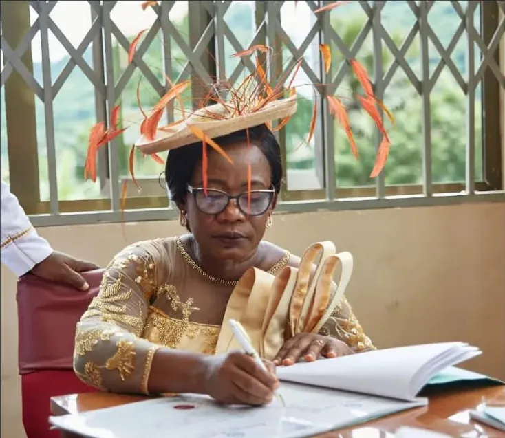 A wedding guest wearing a wide gold fascinator trimmed with orange feathers, signing the register.